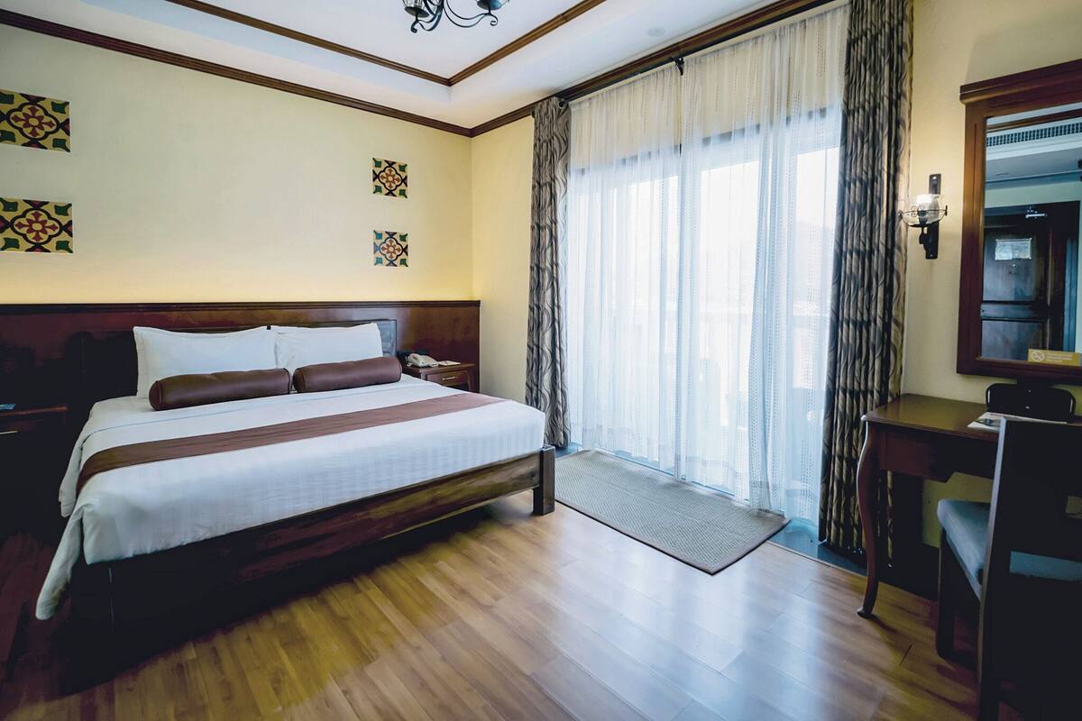 A hotel room with a large bed, a desk, a chair, a mirror, and a window with curtains.