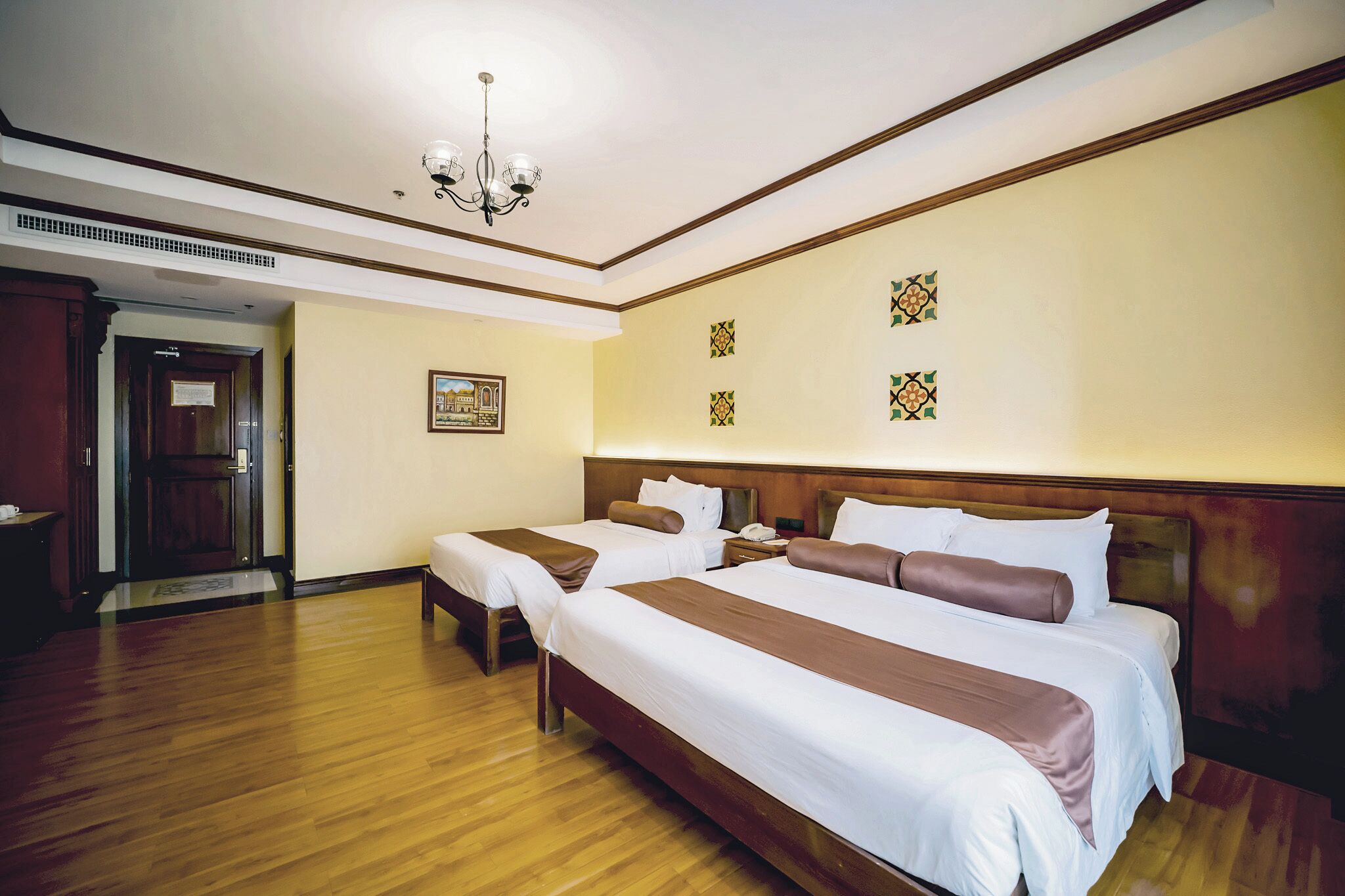 A hotel room with two beds, a wooden floor, a chandelier, and a framed picture on the wall.