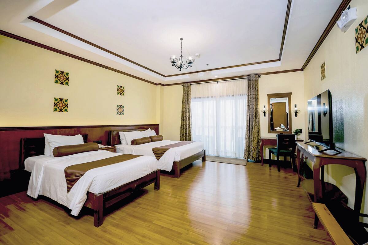A hotel room with two beds, a desk, a television, and a chandelier.