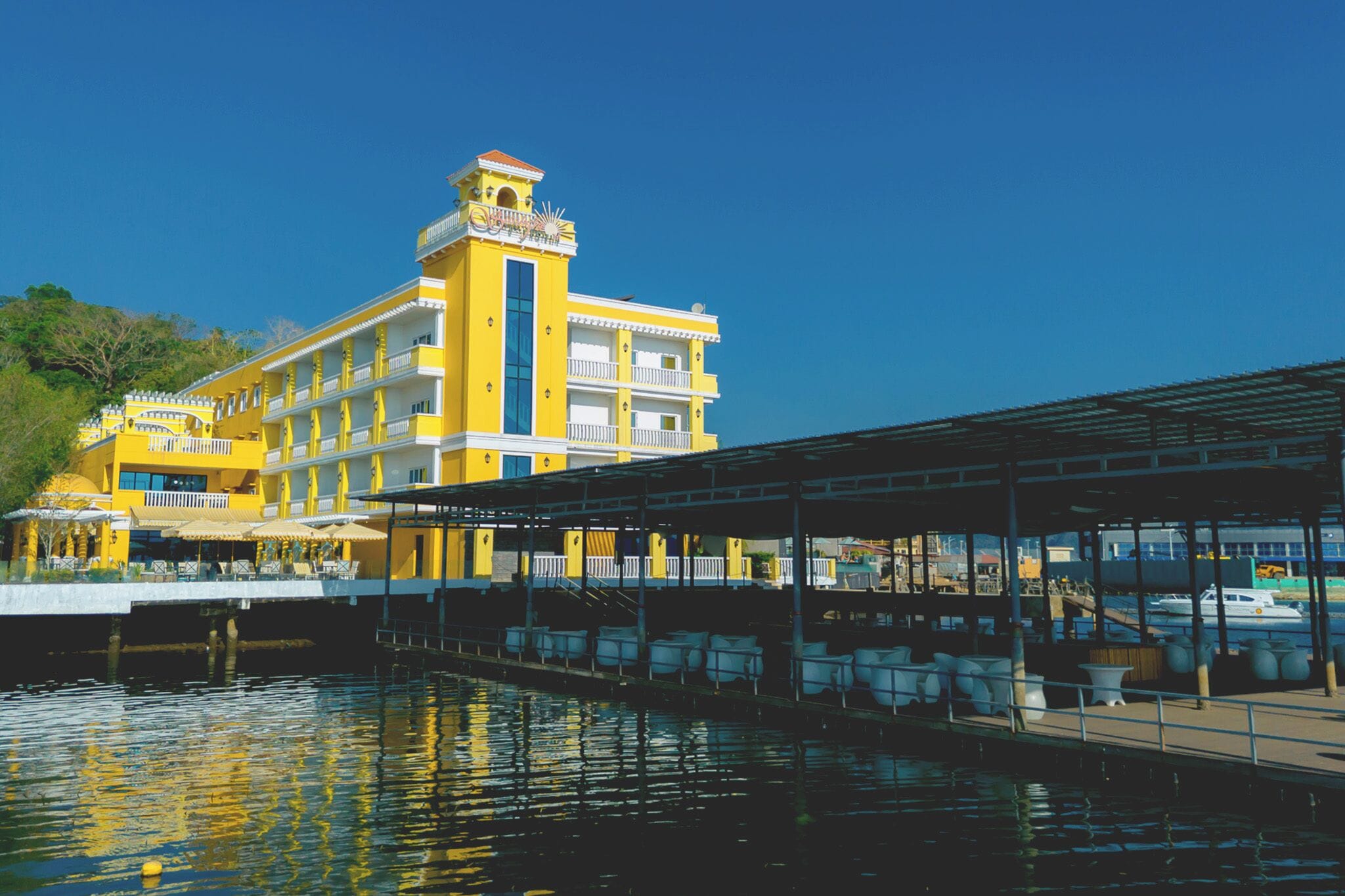 A yellow building with a balcony, a covered area with chairs, and a body of water in front.