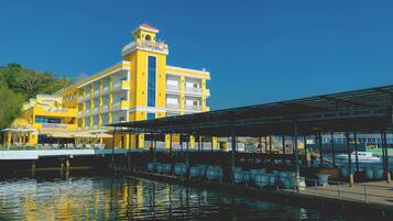 A yellow building with a balcony, a covered area with chairs, and a body of water in front.