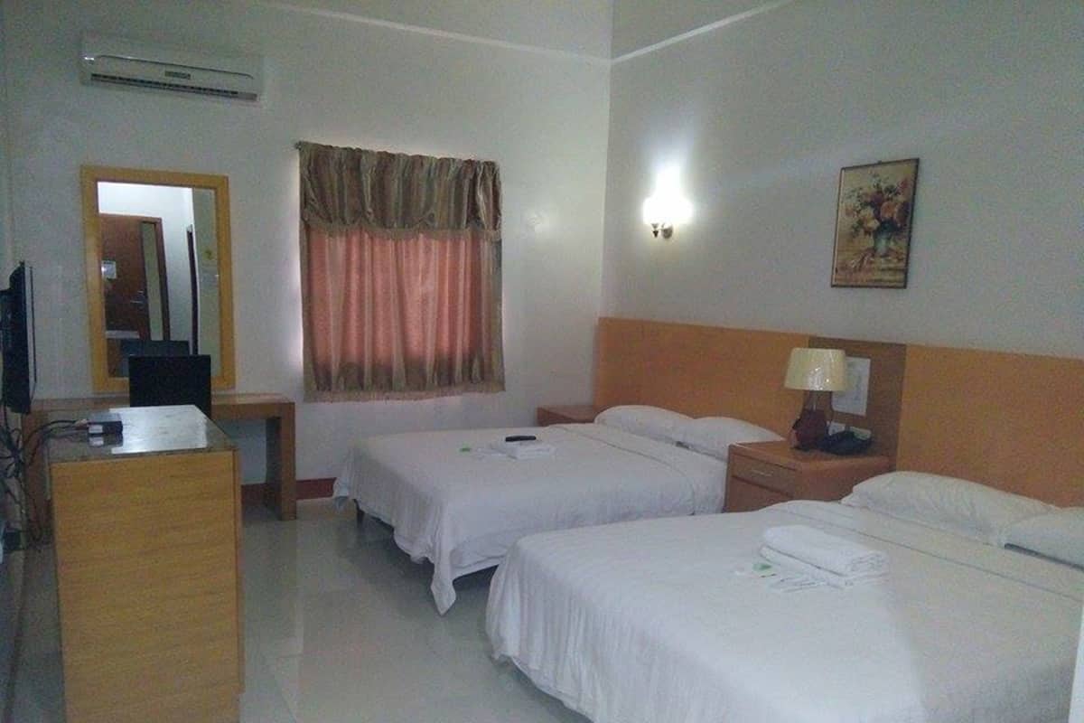 A hotel room with two beds, a desk, a chair, a mirror, a window with curtains, and a wall-mounted lamp.