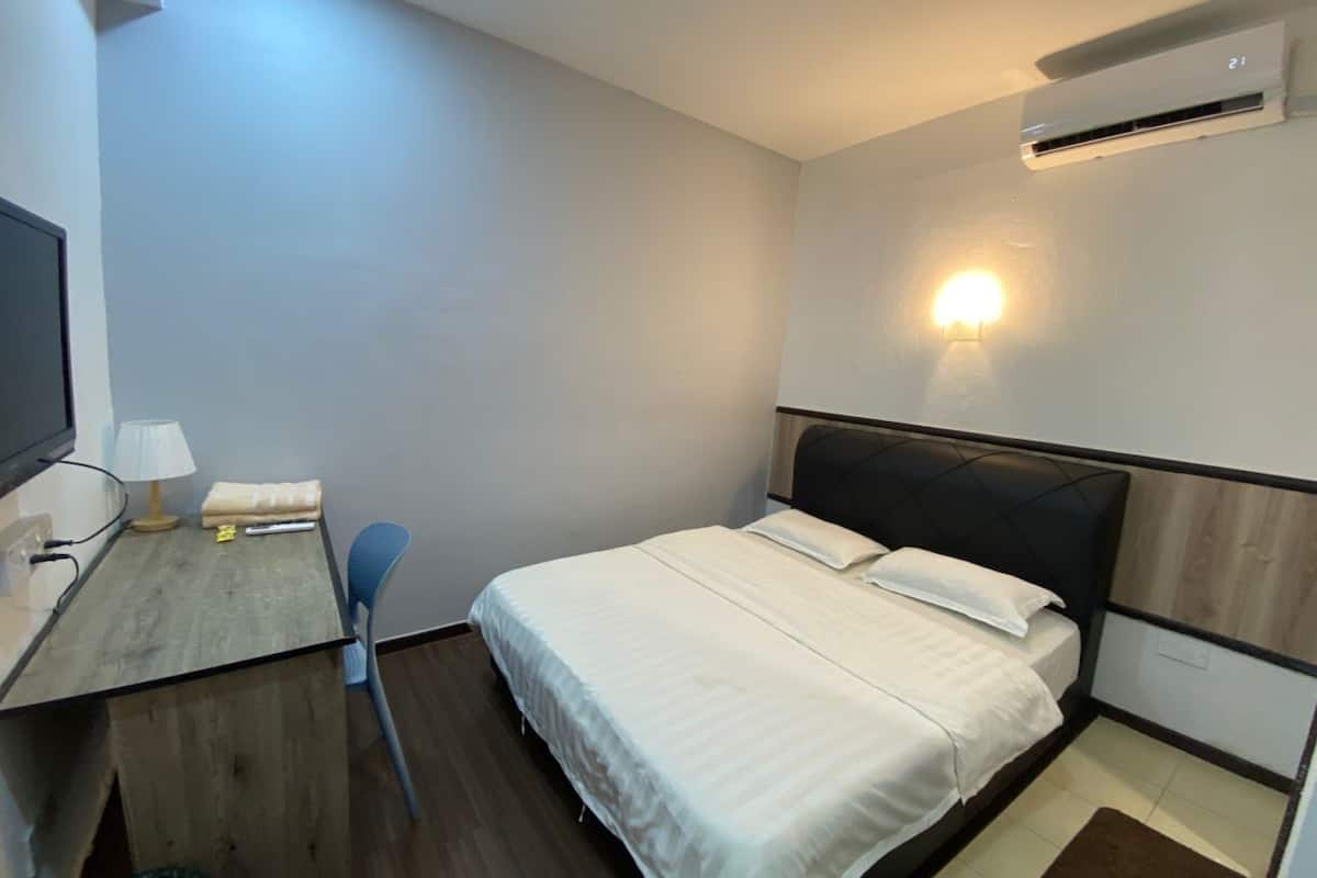 A compact hotel room with a bed, a desk with a lamp, a chair, and a wall-mounted air conditioner.
