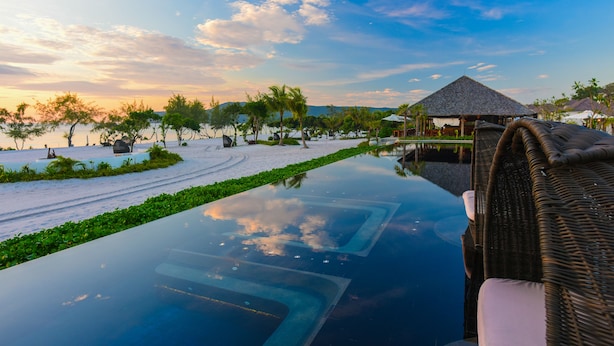 A serene infinity pool with a clear view of the beach and palm trees, surrounded by lush greenery and a thatched-roof structure. - The Royal Sands Koh Rong (Koh Rong)