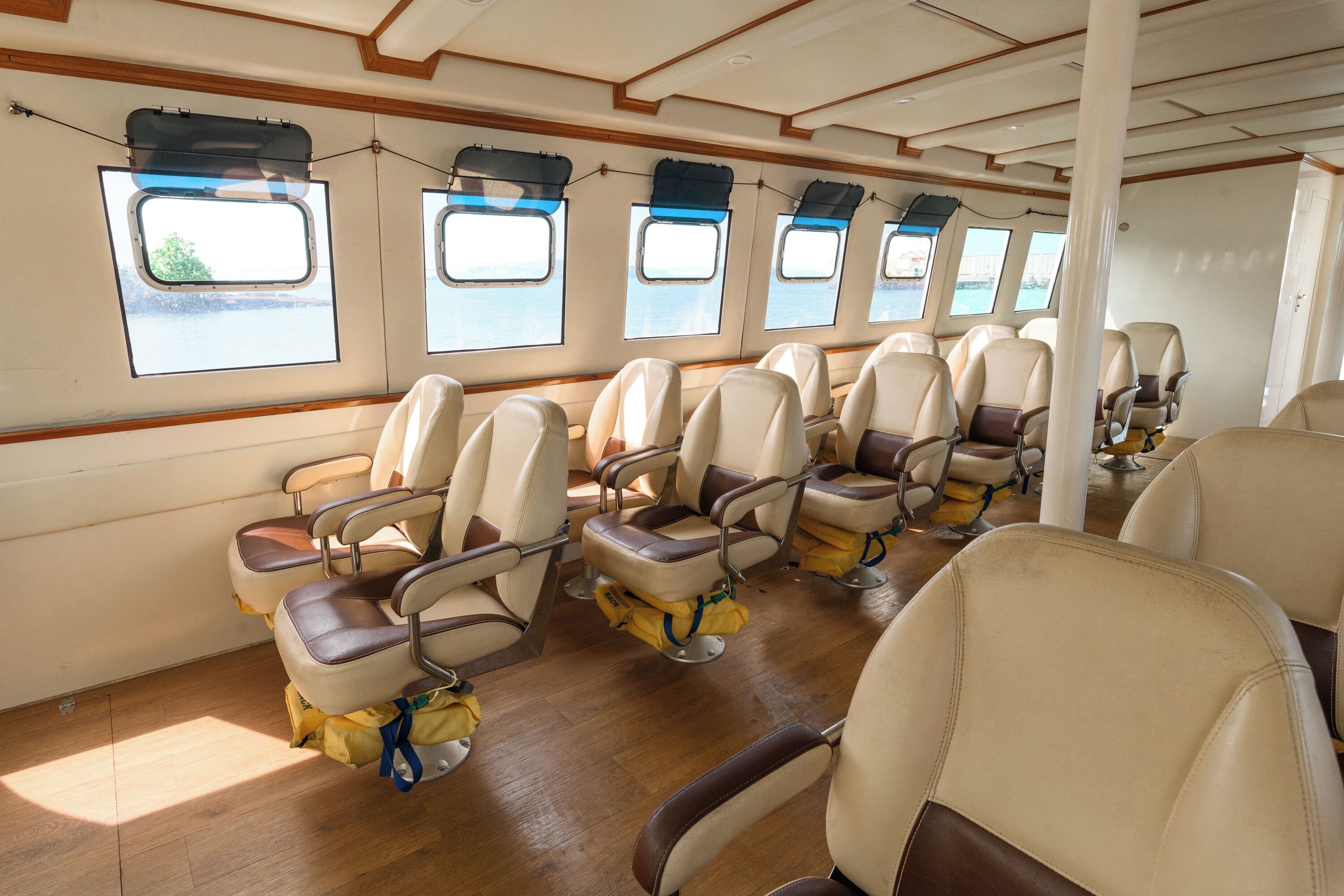 A row of seats with overhead luggage racks, windows with views of the water, and a wooden floor.