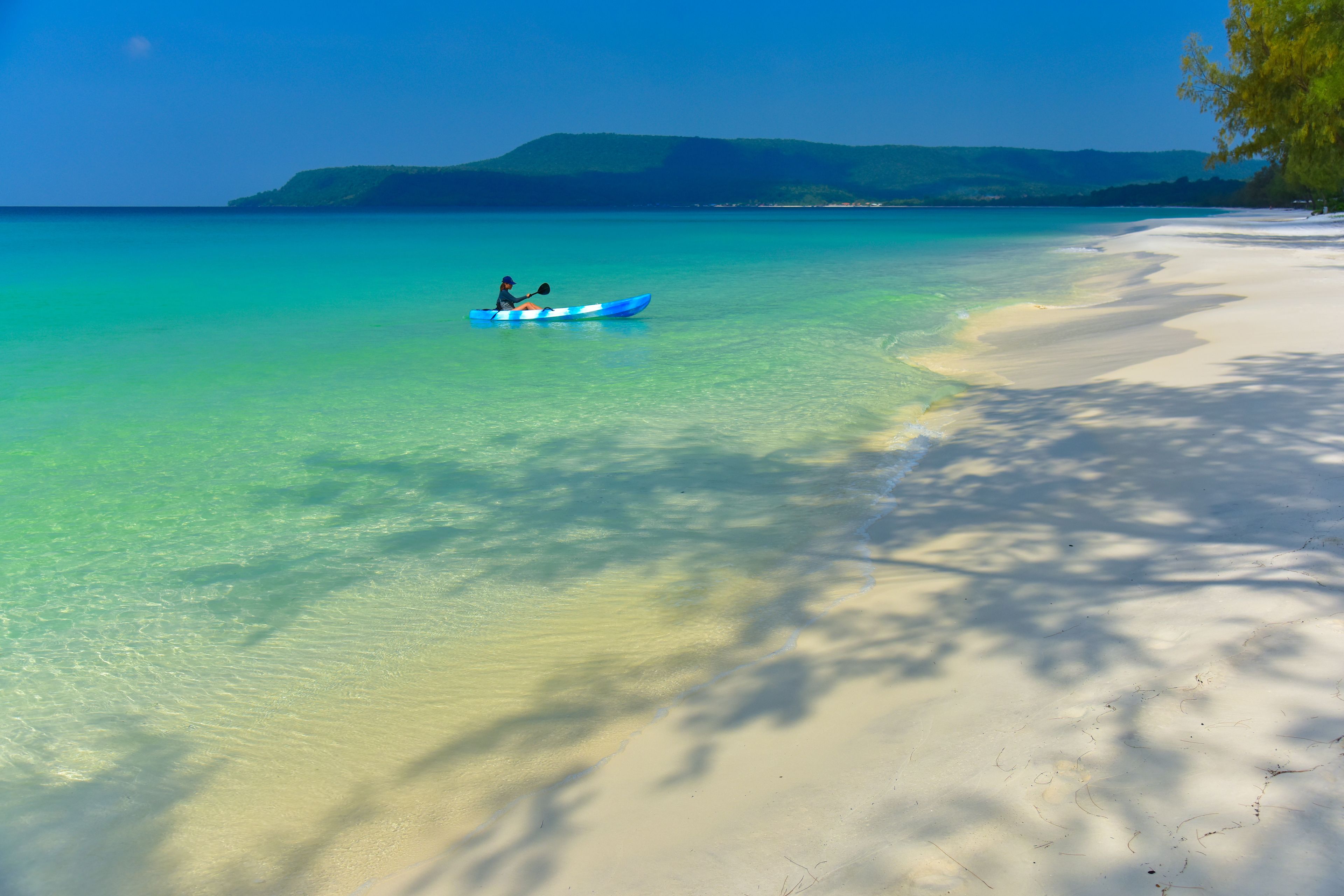 Un kayakista en un mar turquesa y transparente, con una playa arenosa y colinas a lo lejos.