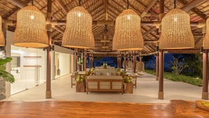 A spacious indoor area with a high ceiling, wooden beams, and hanging wicker lamps. There is a seating area with a sofa and wooden stools, surrounded by greenery. - The Royal Sands Koh Rong (Koh Rong)