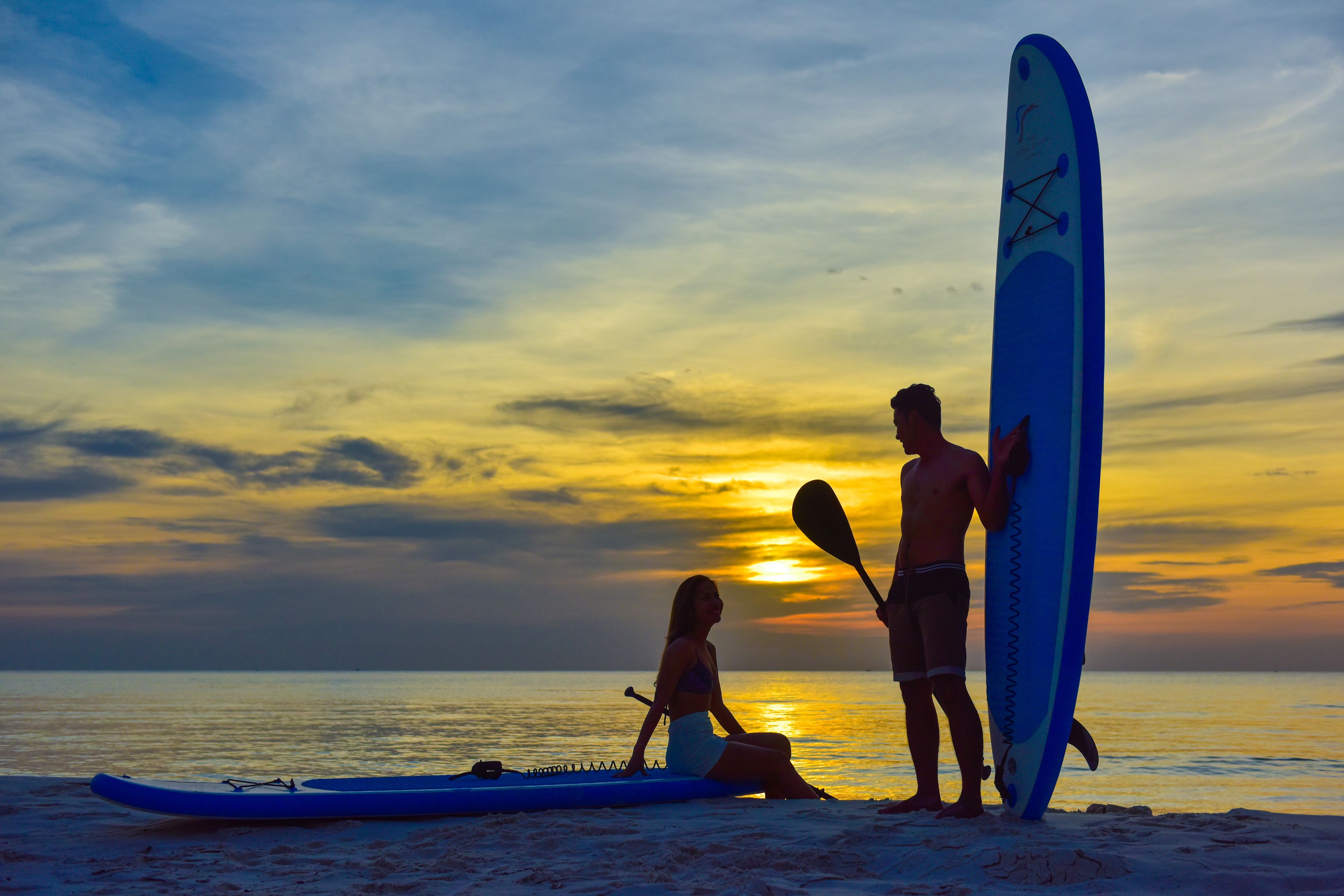 Escena de playa con atardecer, una tabla de surf y un remo.