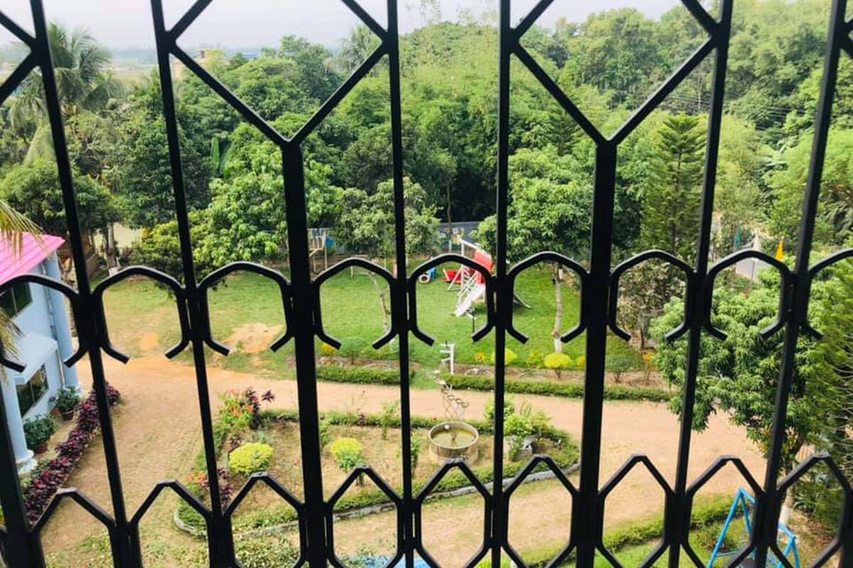 A view through a metal gate of a landscaped garden with a playground, a white building, and a clear sky.