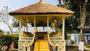 A thatched-roof gazebo with a staircase leading up to a bench area, surrounded by colorful stones and greenery. - Tea Heaven Resort (Sreemangal)
