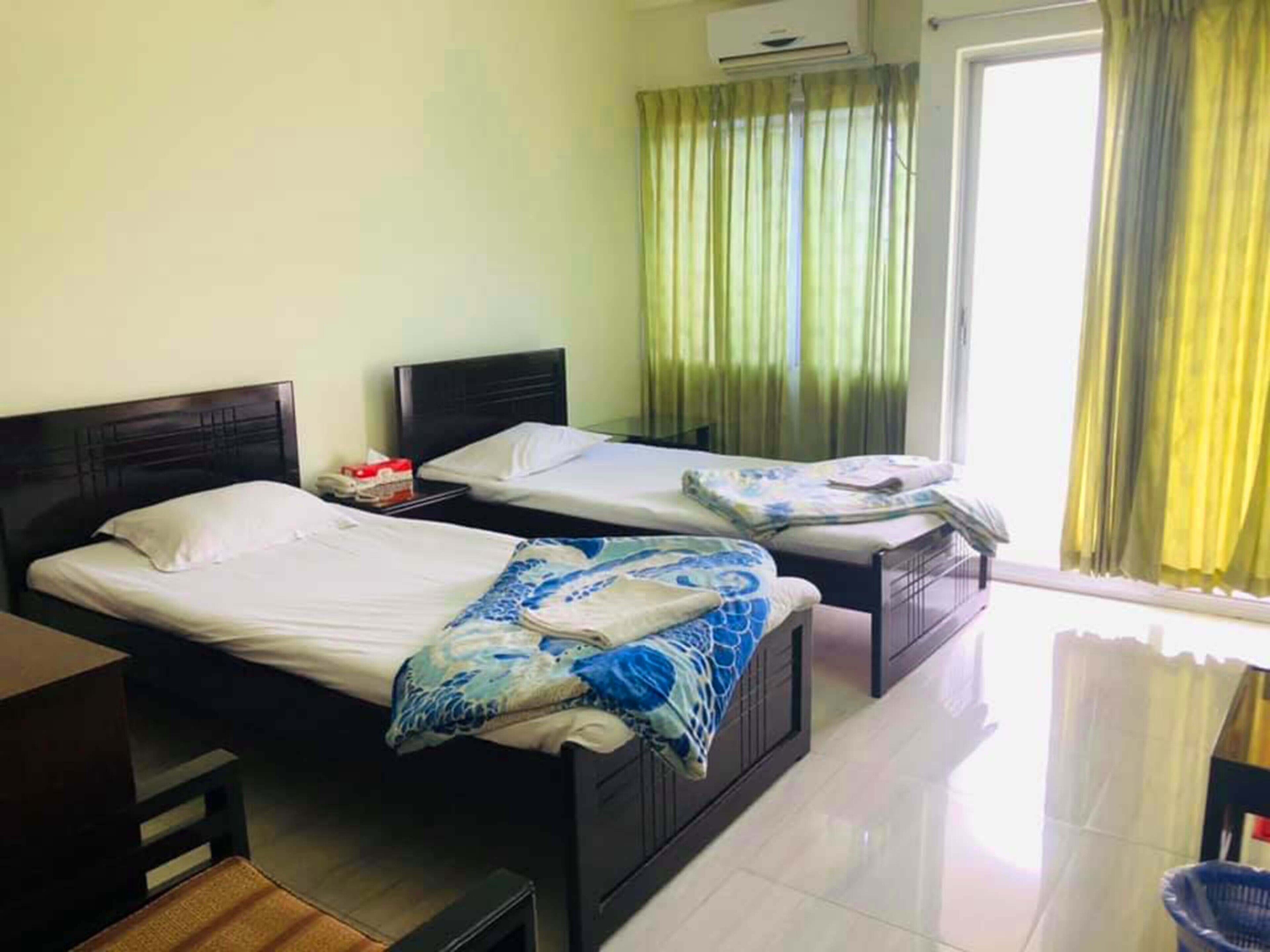 A hotel room with two beds, a desk, and a chair. There is an air conditioning unit on the wall and a window with curtains.