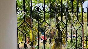 A window with a metal grille, through which a garden with palm trees and a playground is visible. - Tea Heaven Resort (Sreemangal)