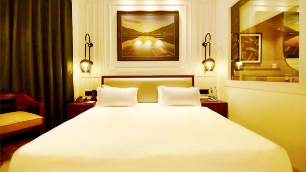 Quarto de hotel com uma cama grande, duas mesas de cabeceira, uma cadeira, um quadro na parede e uma janela com vista.
