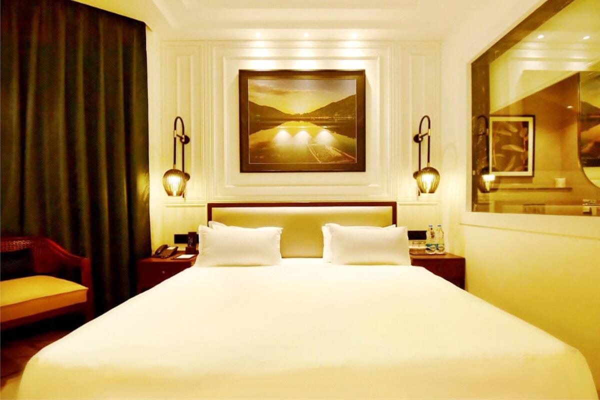 A hotel room with a large bed, two bedside tables, a chair, a painting on the wall, and a window with a view.