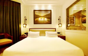 A hotel room with a large bed, two bedside tables, a chair, a painting on the wall, and a window with a view. - Hotel Rani Castle (New Delhi)