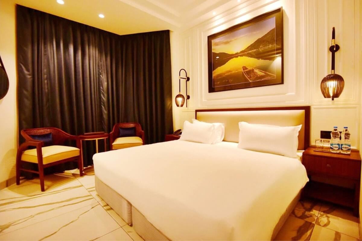 A hotel room with a large bed, two chairs, a nightstand with water bottles, and a painting on the wall.