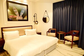 A hotel room with a large bed, two chairs, a small table, a wall-mounted mirror, and a painting of boats. - Hotel Rani Castle (New Delhi)