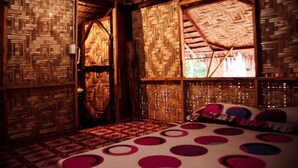 Um quarto com telhado de palha, paredes de bambu trançado, piso estampado e uma cama com almofadas circulares.
