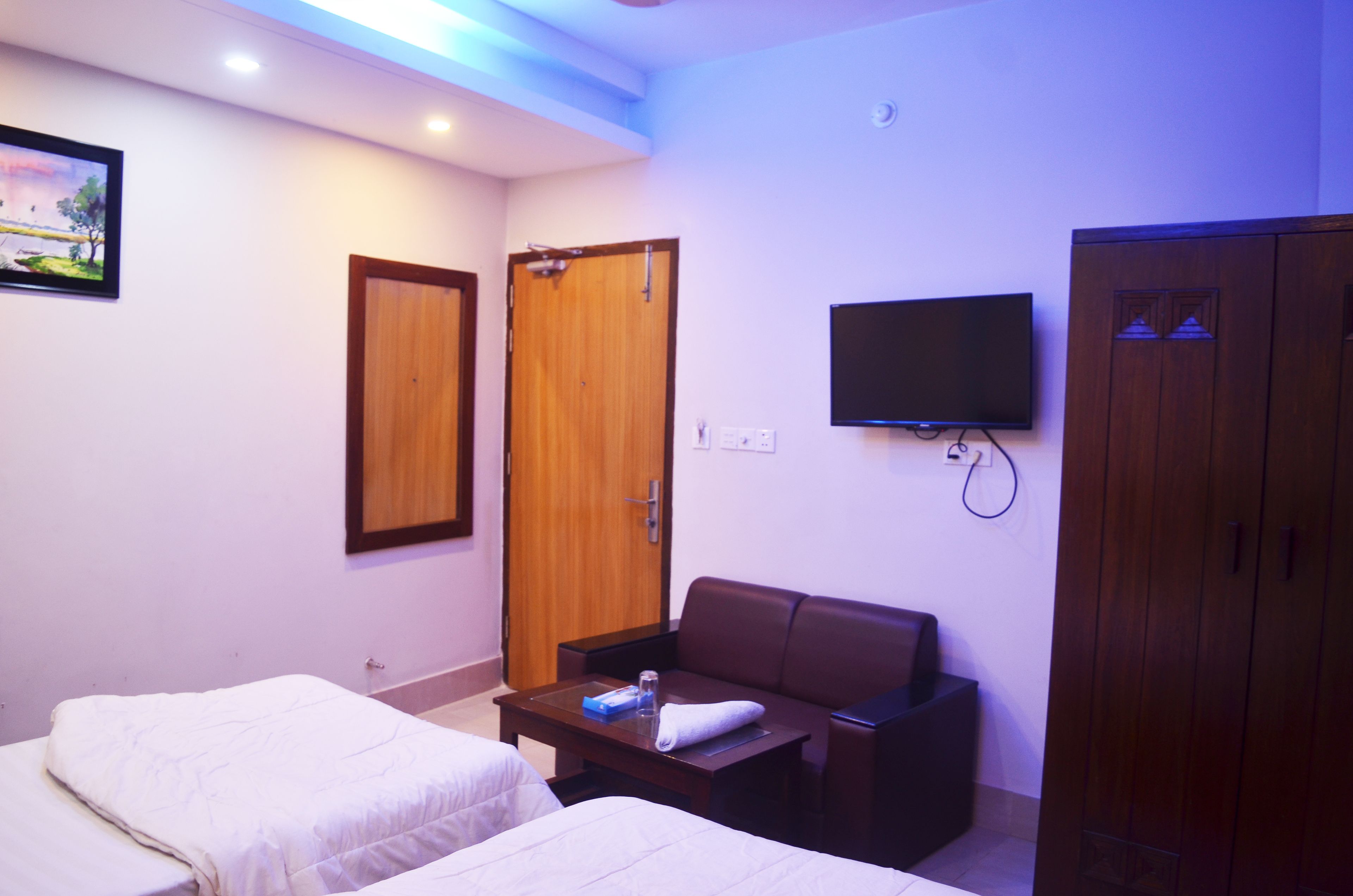 A hotel room with two beds, a TV, a wooden wardrobe, and a small table with a bottle and glasses.