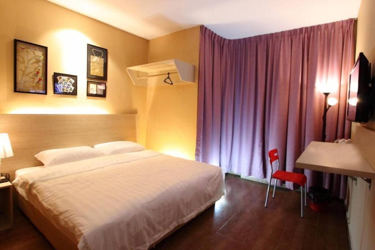 A hotel room with a bed, bedside table, lamp, wall-mounted TV, and a window with curtains.