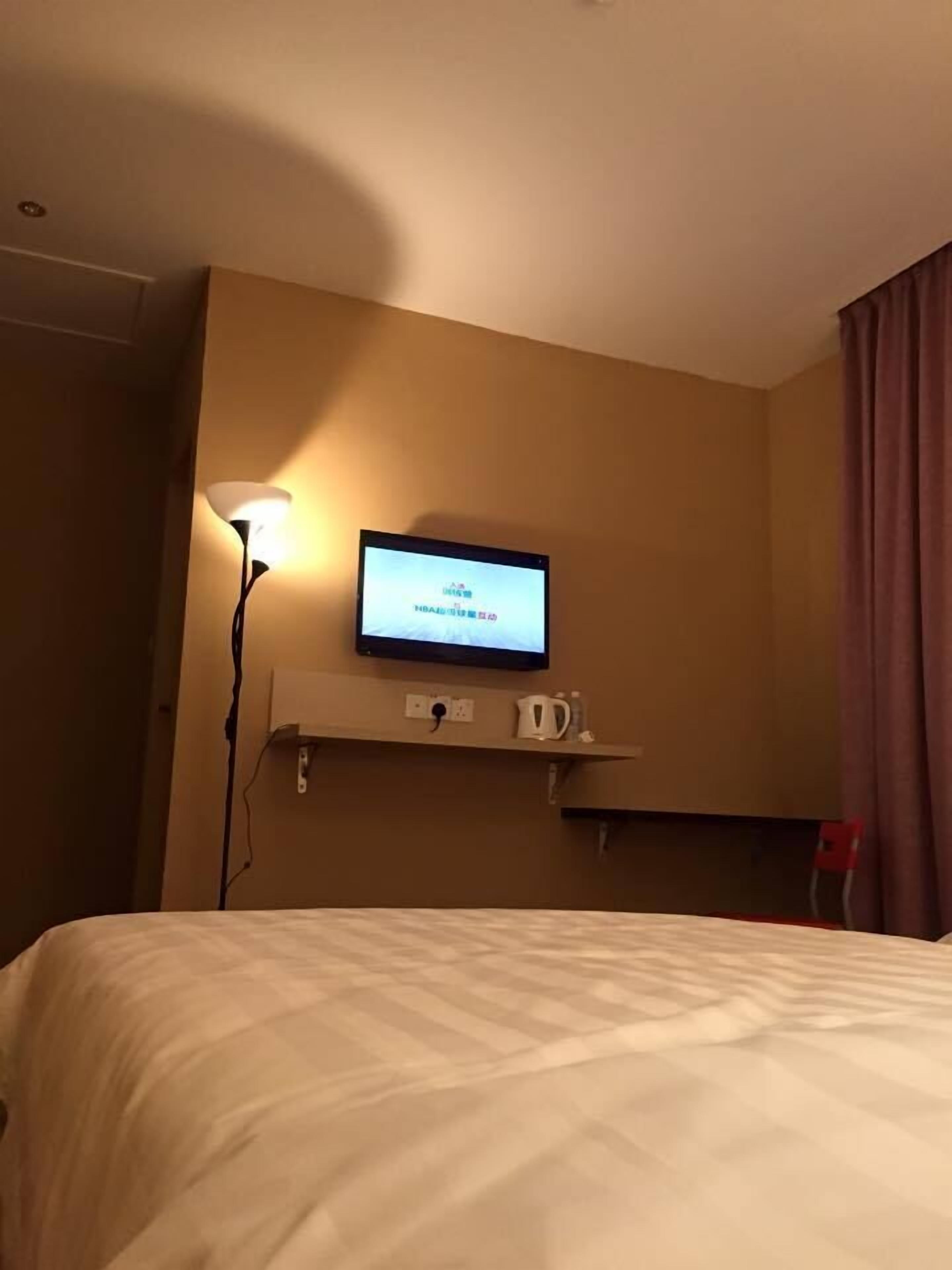 A hotel room with a bed, a mounted television displaying text, a bedside table with a cup and a kettle, and a lamp.