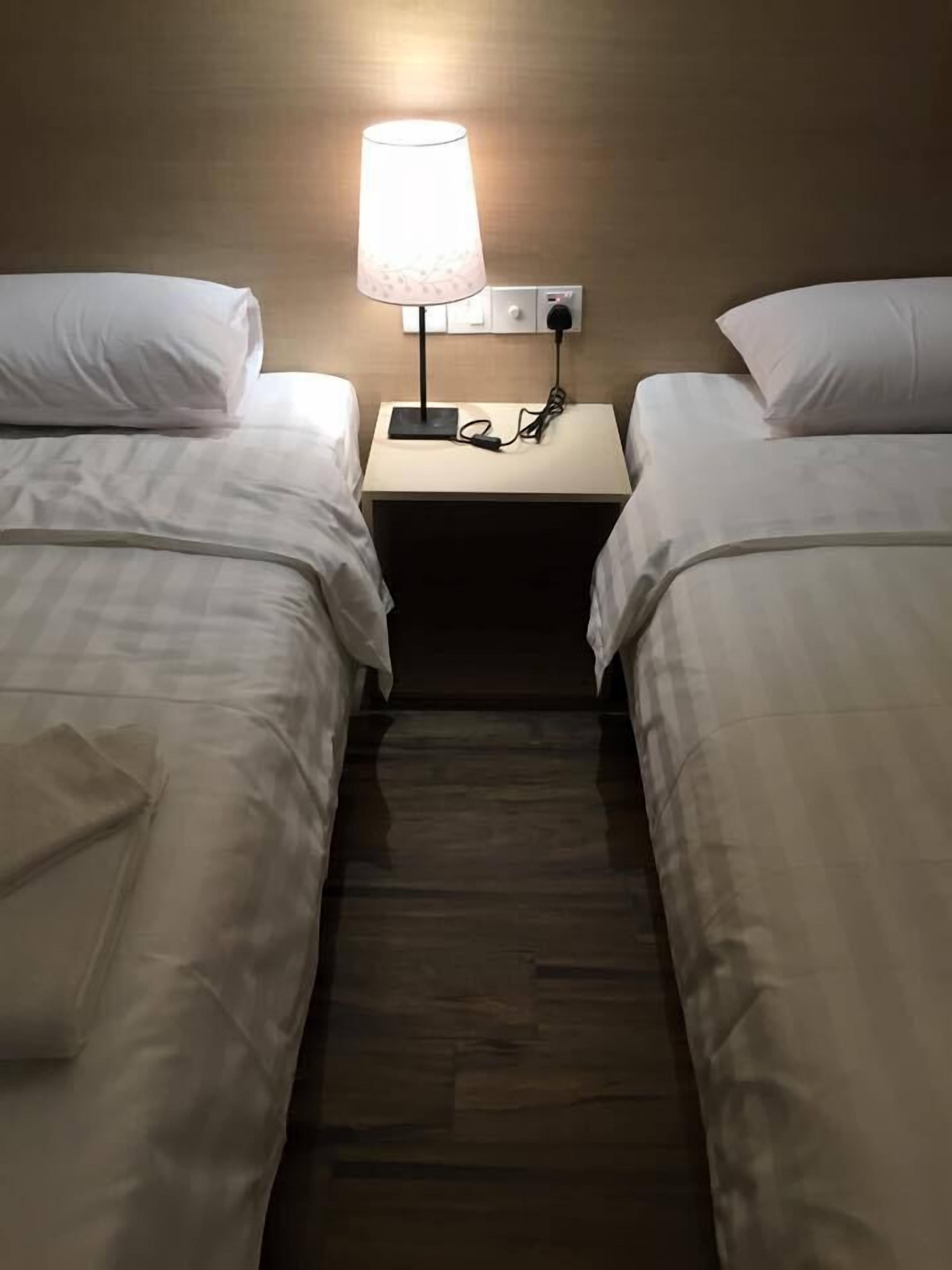 A hotel room with two single beds, a nightstand with a lamp, and a power outlet.
