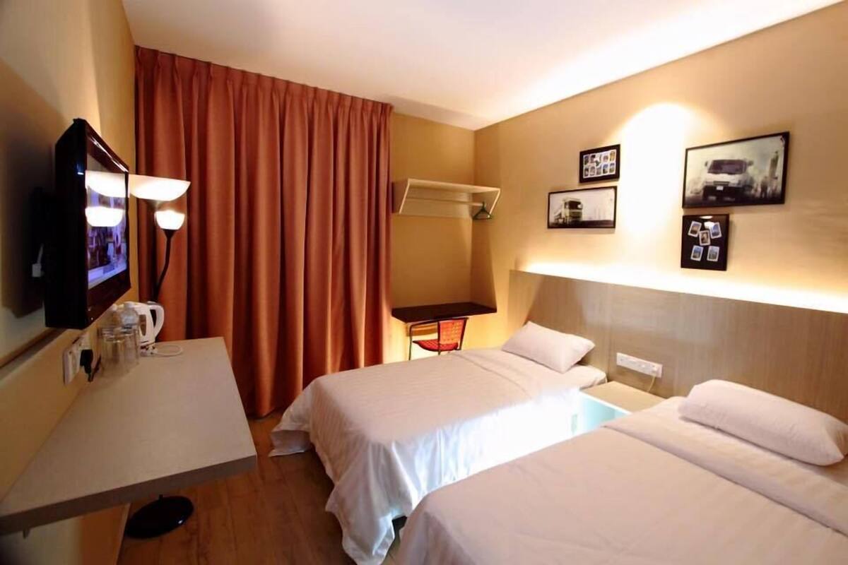 A hotel room with two beds, a desk with a lamp, a TV, and a wall-mounted shelf with pictures.