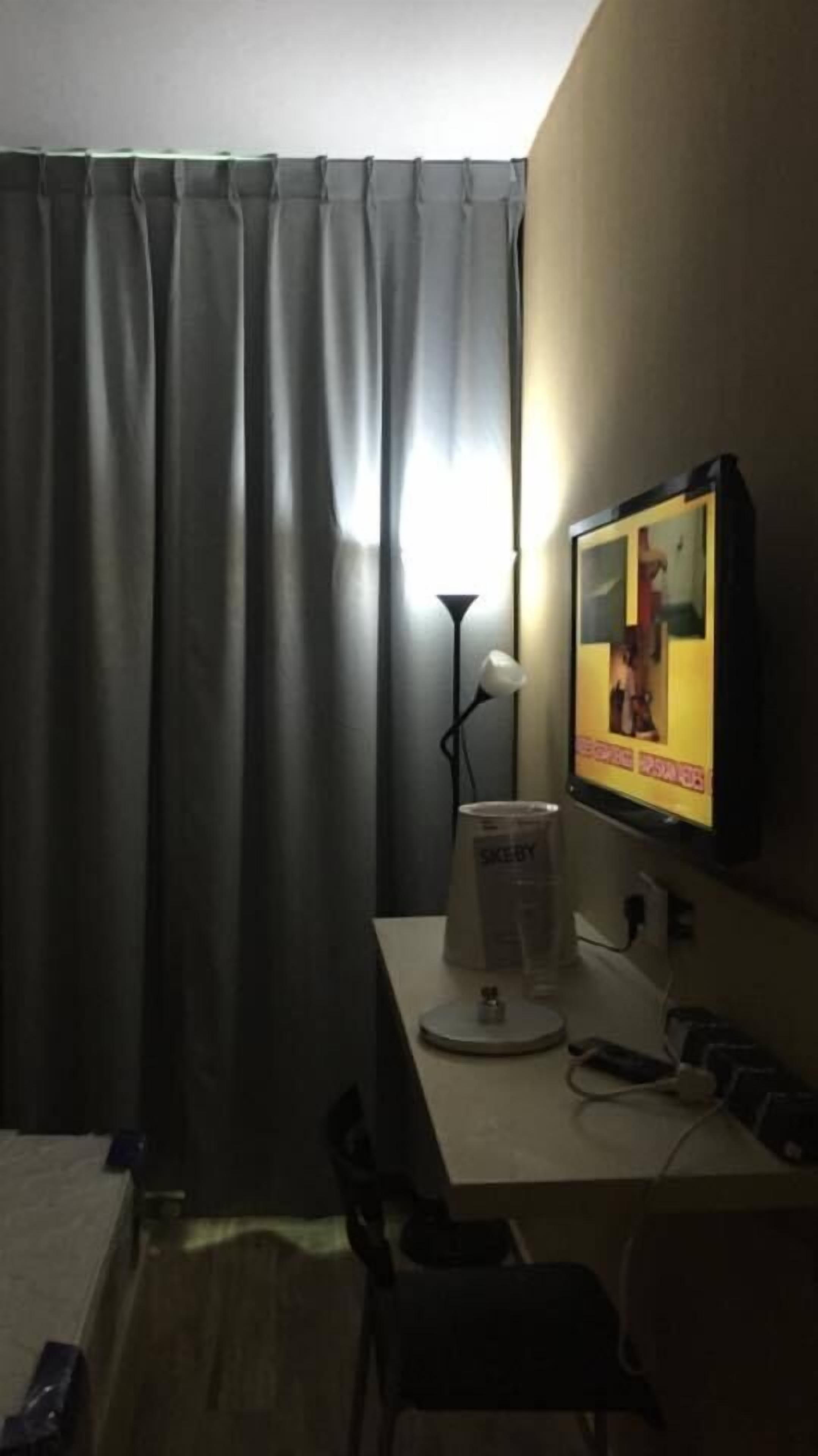 A hotel room with a bed, a desk with a computer, a lamp, and a TV displaying a person.