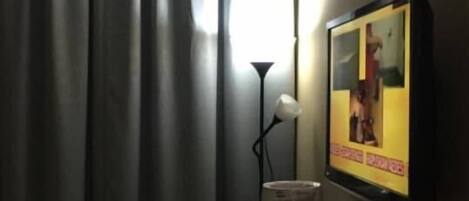 A hotel room with a bed, a desk with a computer, a lamp, and a TV displaying a person.