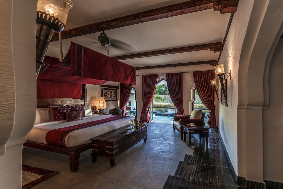 A bedroom with a canopy bed, a sitting area with a table and chairs, and a view of a garden through a window.