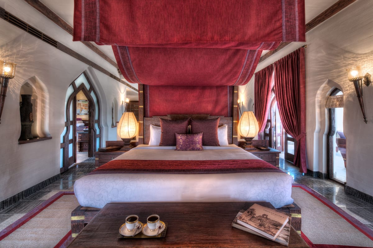 A luxurious hotel room with a large bed, ornate headboard, and a red draped ceiling. There are two bedside lamps, a wooden nightstand, and a pair of slippers on the floor.