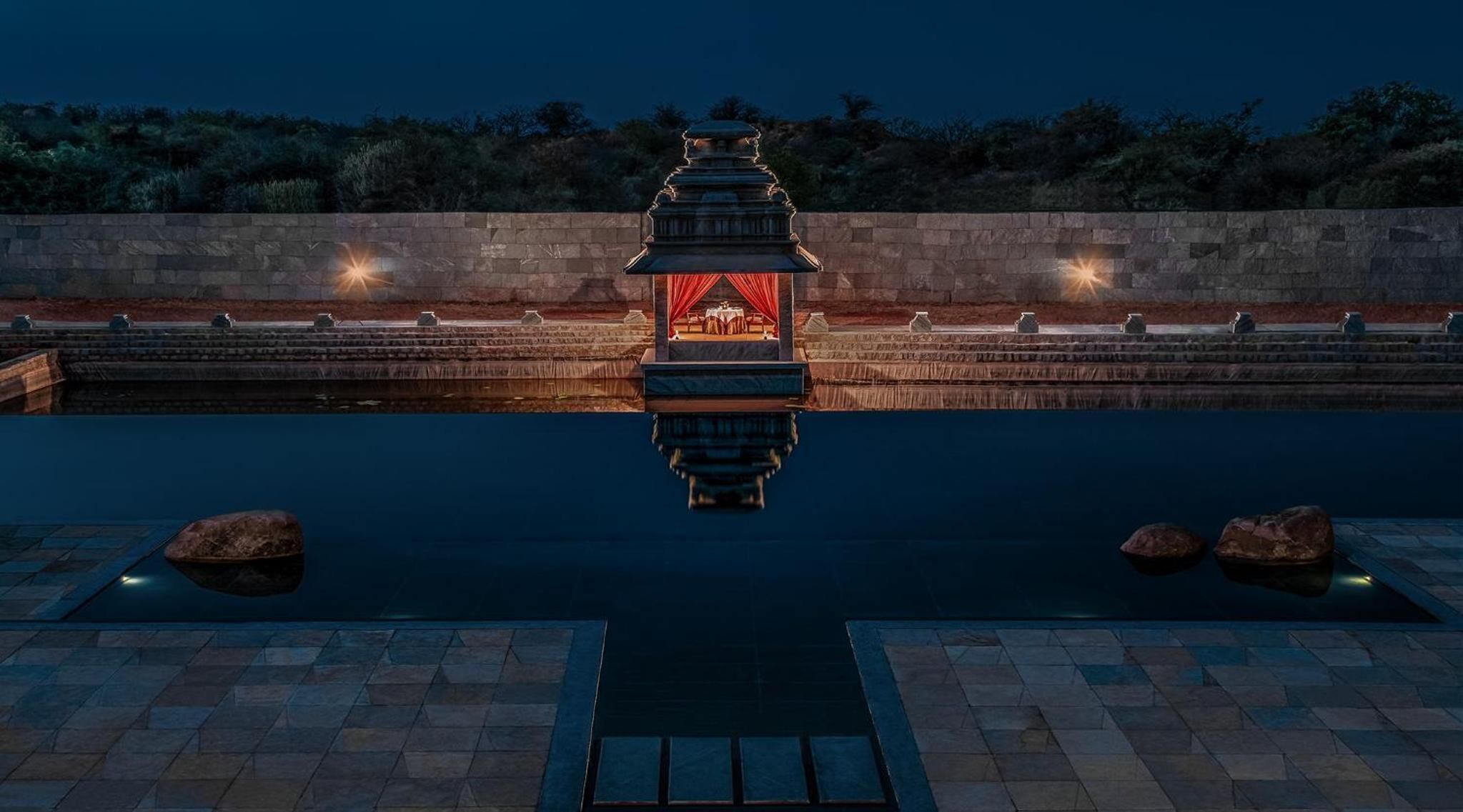 A serene pool area with a traditional pagoda-style structure in the center, surrounded by stone walls and a calm water surface.