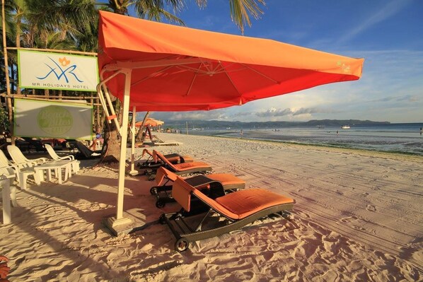 A beachside hotel with lounge chairs and umbrellas, a sign for 'Mr. Holidays Hotel', and a view of the ocean.