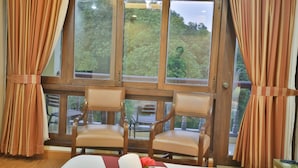 Une pièce avec une grande fenêtre, deux chaises en bois, un lit recouvert d’une couette blanche et une fleur rouge posée sur le lit.