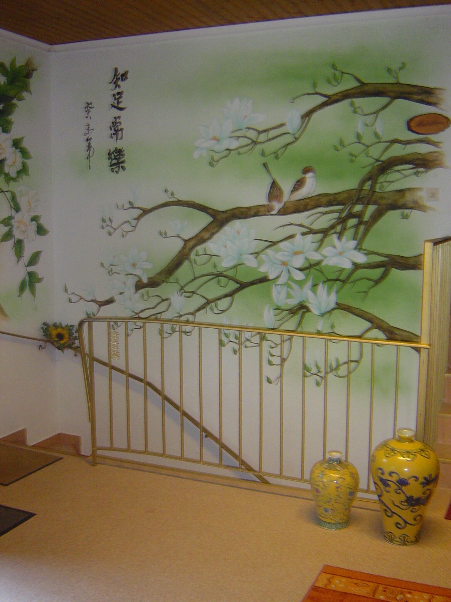 A room with a mural of a tree with white flowers and a bird, a metal gate, and two yellow vases.