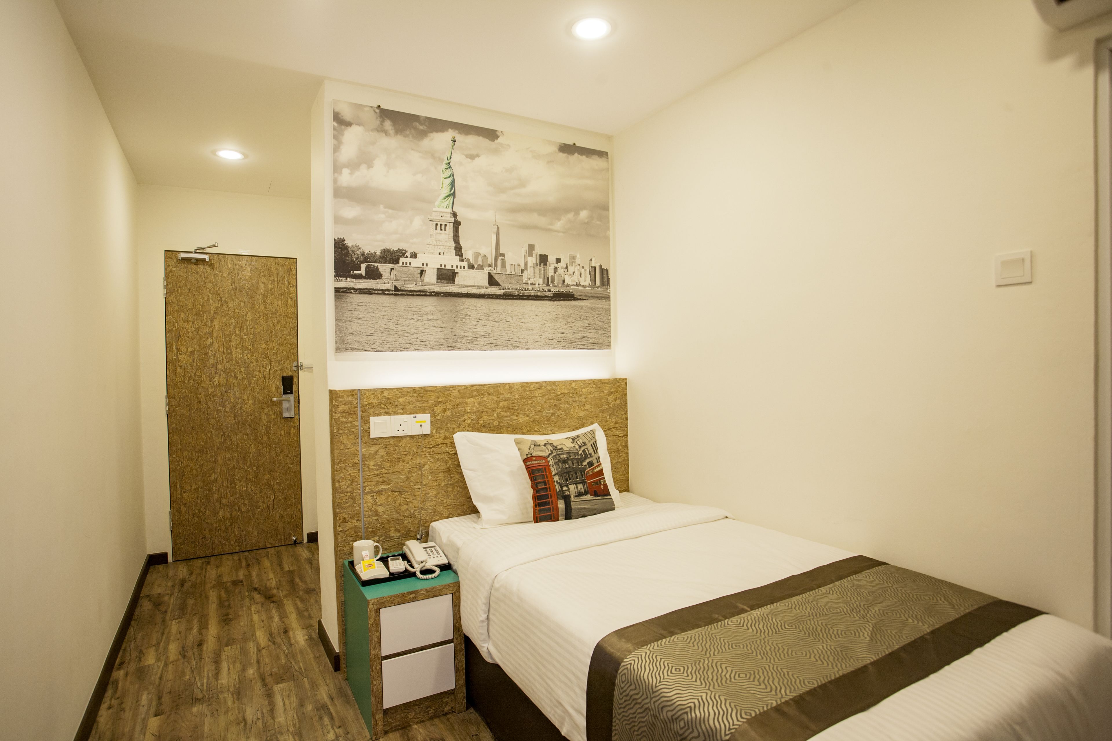 A hotel room with a bed, a nightstand with a phone, a wall-mounted light, and a door.