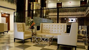 Um hall com chão quadriculado, um sofá branco, duas poltronas e uma mesa de centro de vidro com um arranjo floral.