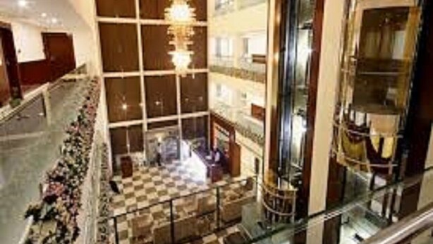 Um hall amplo com piso xadrez, um lustre central e um guarda-corpo de vidro com vista para o espaço.
