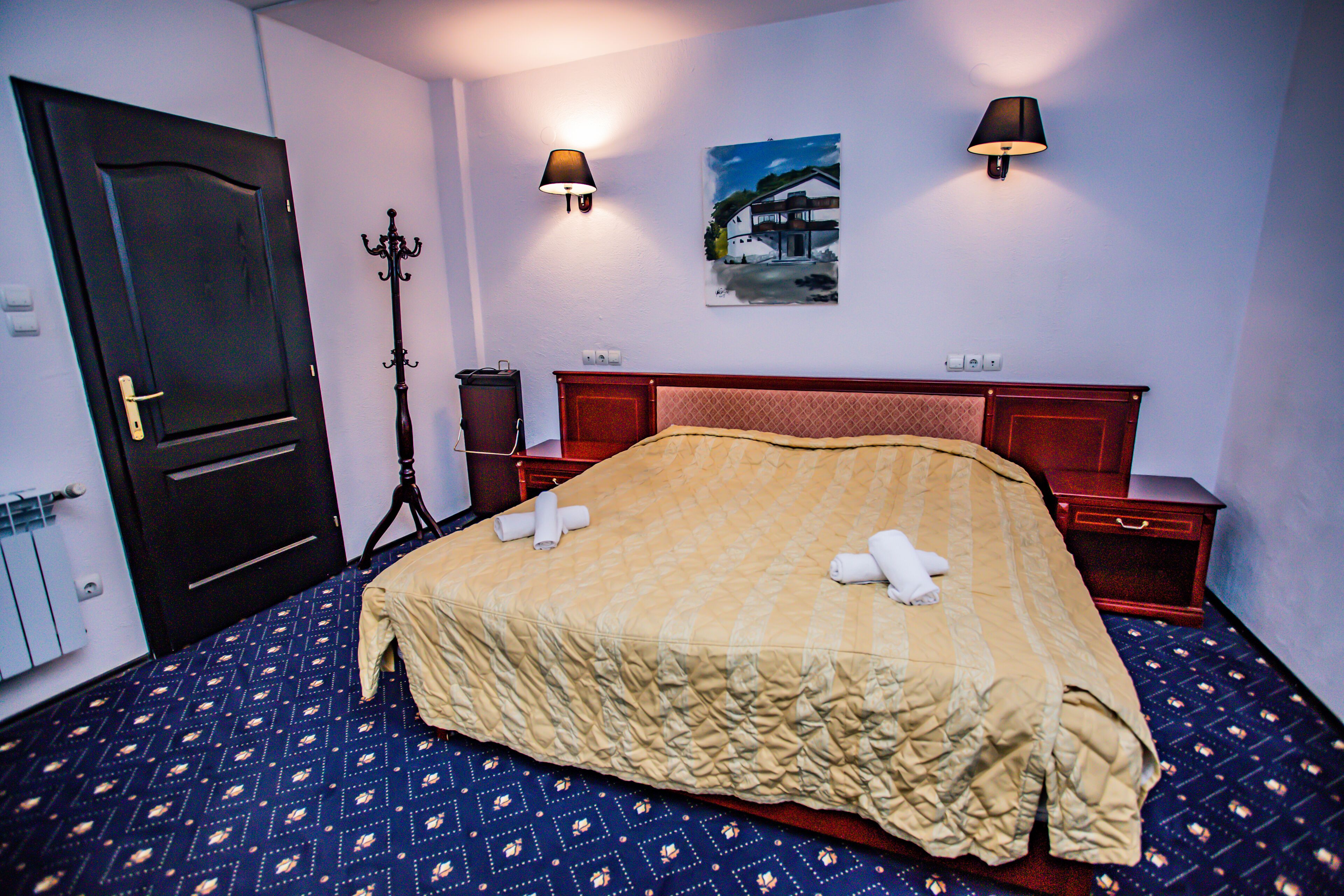 A hotel room with a large bed, two bedside lamps, a painting on the wall, and a coat rack.