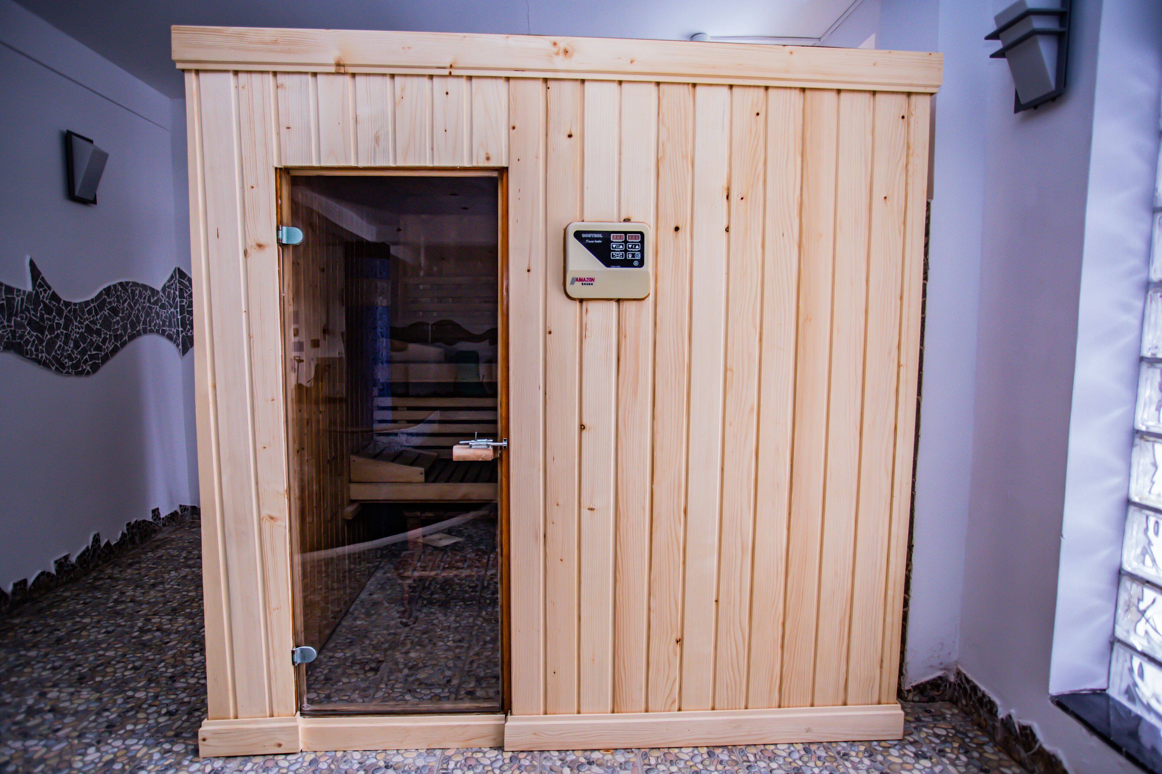 A wooden sauna with a control panel, a glass door, and a pebble floor.