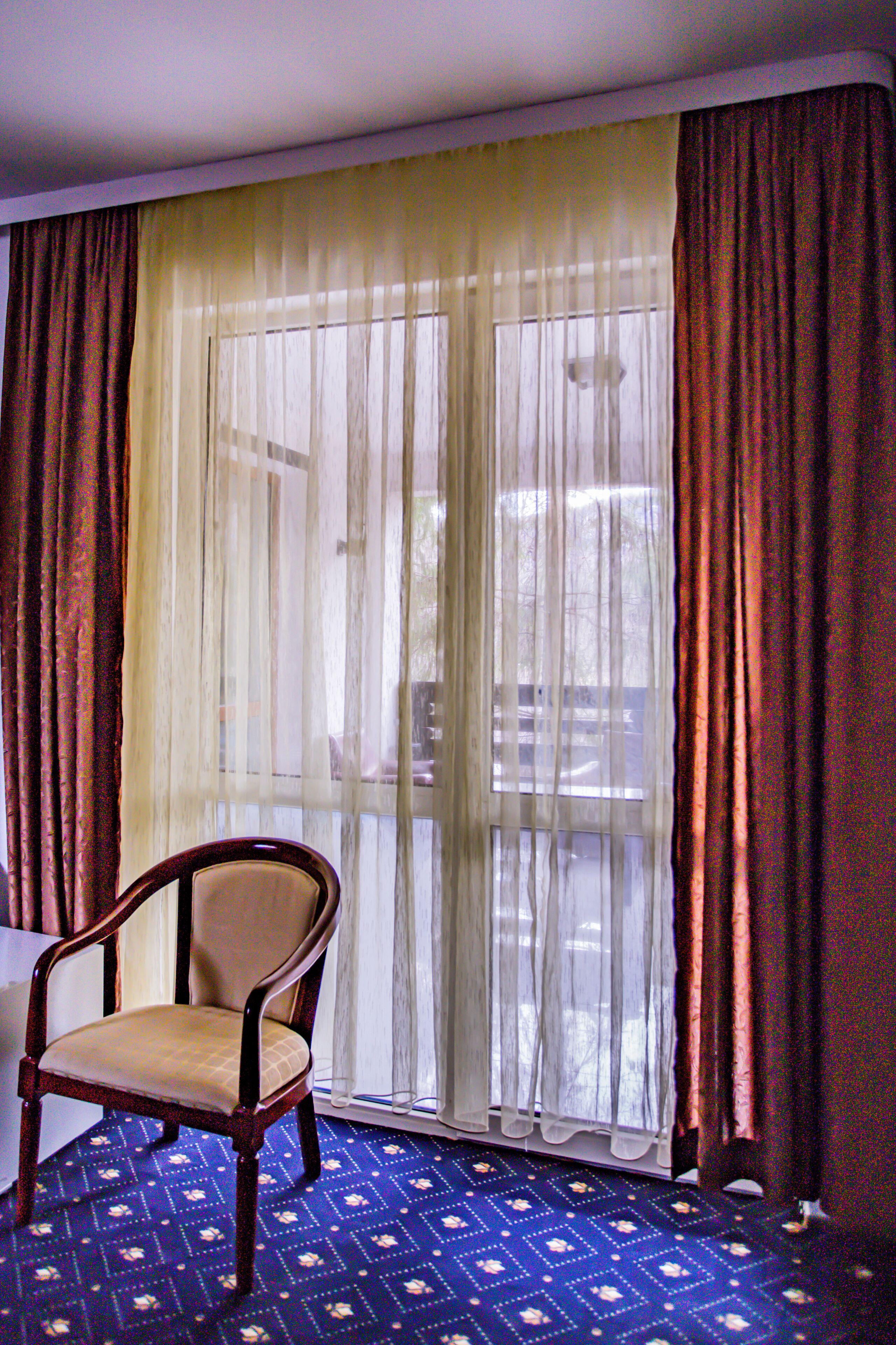 Ein Zimmer mit einem Sessel, einem großen Fenster mit durchsichtigen Vorhängen und einem gemusterten Teppich.