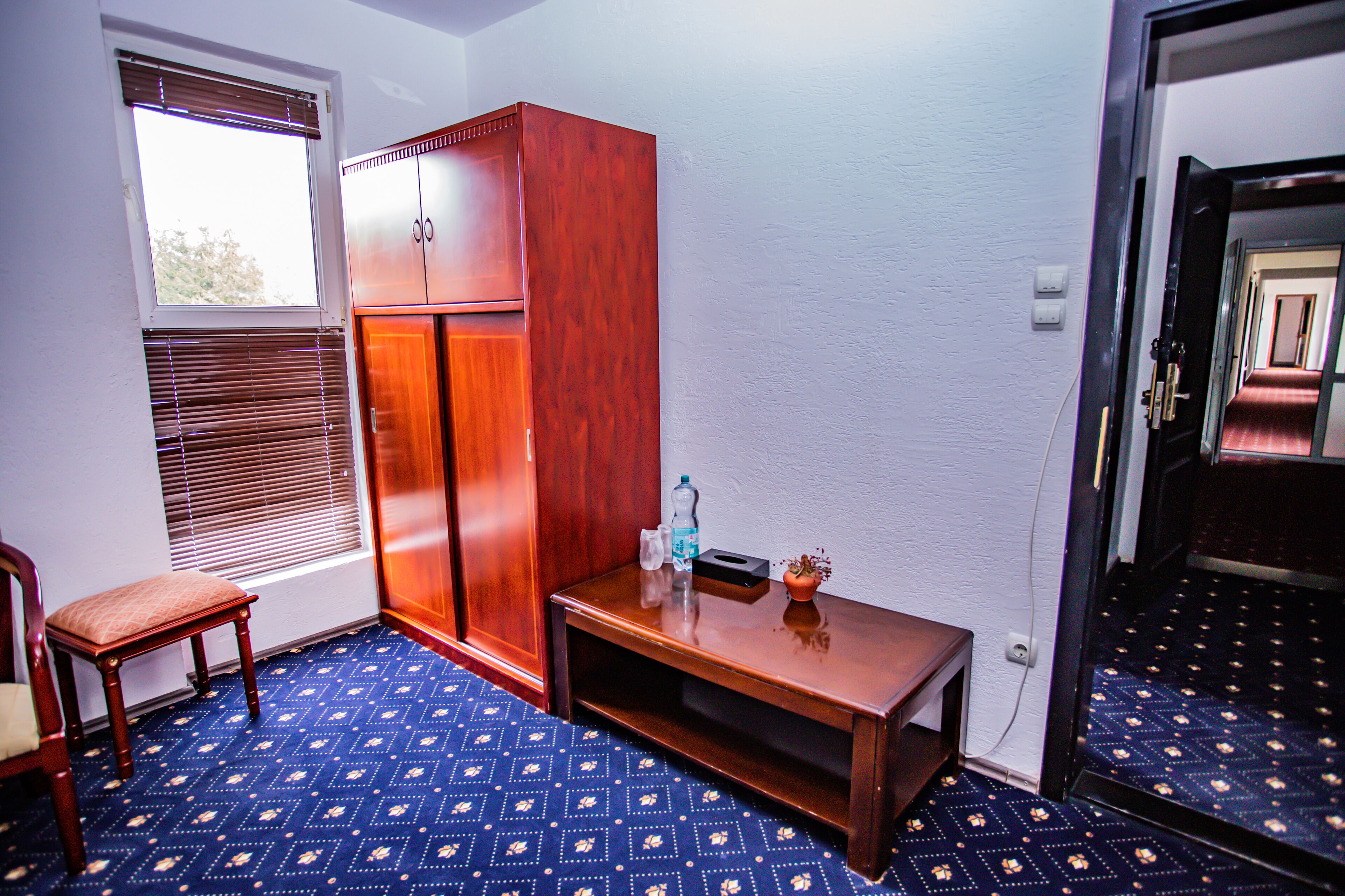 Una habitación con un armario de madera, una mesita con una botella y vasos, una silla y una alfombra estampada.