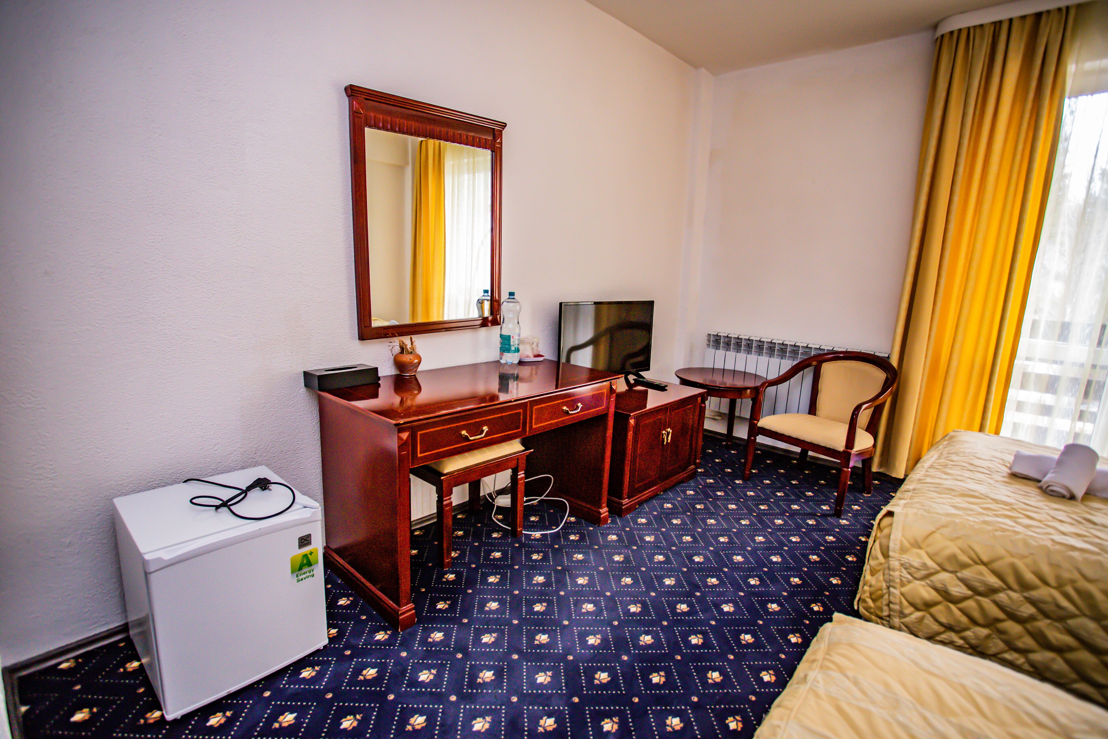 A hotel room with a bed, a desk with a mirror, a television, a chair, and a small refrigerator.