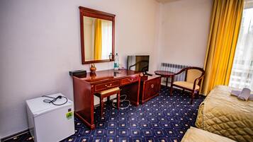 A hotel room with a bed, a desk with a mirror, a television, a chair, and a small refrigerator.