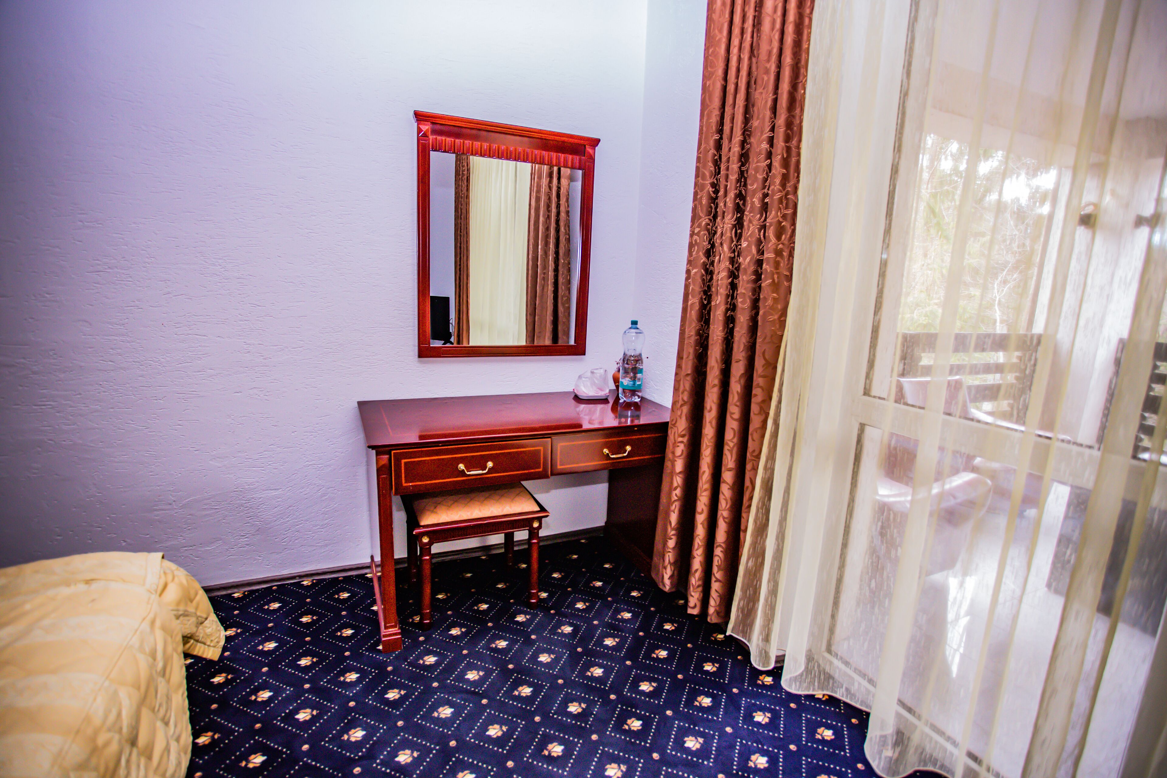 Quarto de hotel com cama, escrivaninha de madeira com gavetas, pequeno banco, espelho, garrafa de água e janela com cortinas.