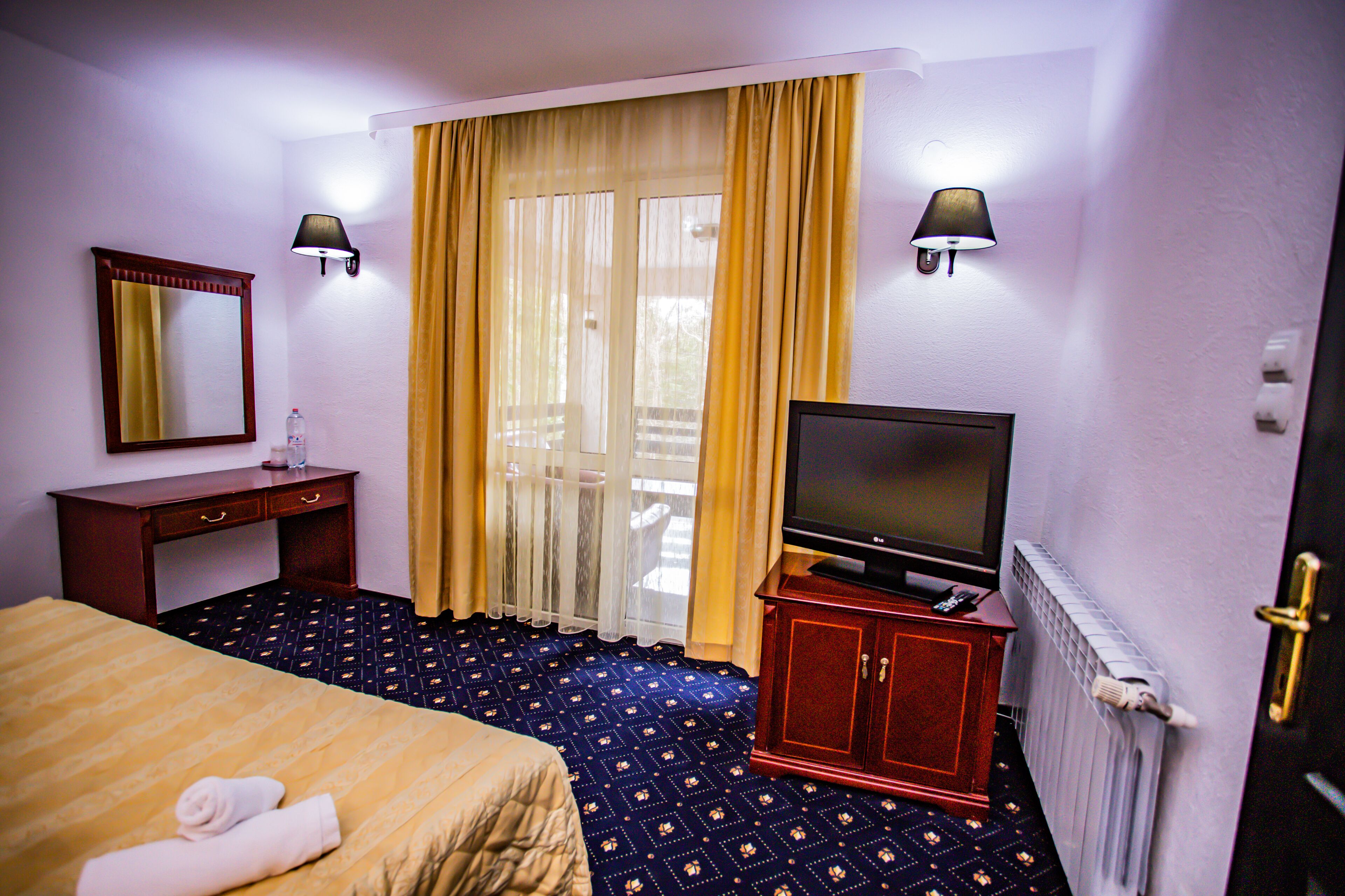 A hotel room with a bed, a TV on a stand, a desk with a lamp, and a window with curtains.