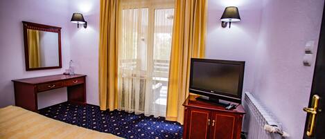 A hotel room with a bed, a TV on a stand, a desk with a lamp, and a window with curtains.