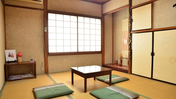Una habitación con esteras de tatami, una mesa de madera, cojines verdes, una ventana y puertas correderas tradicionales.
