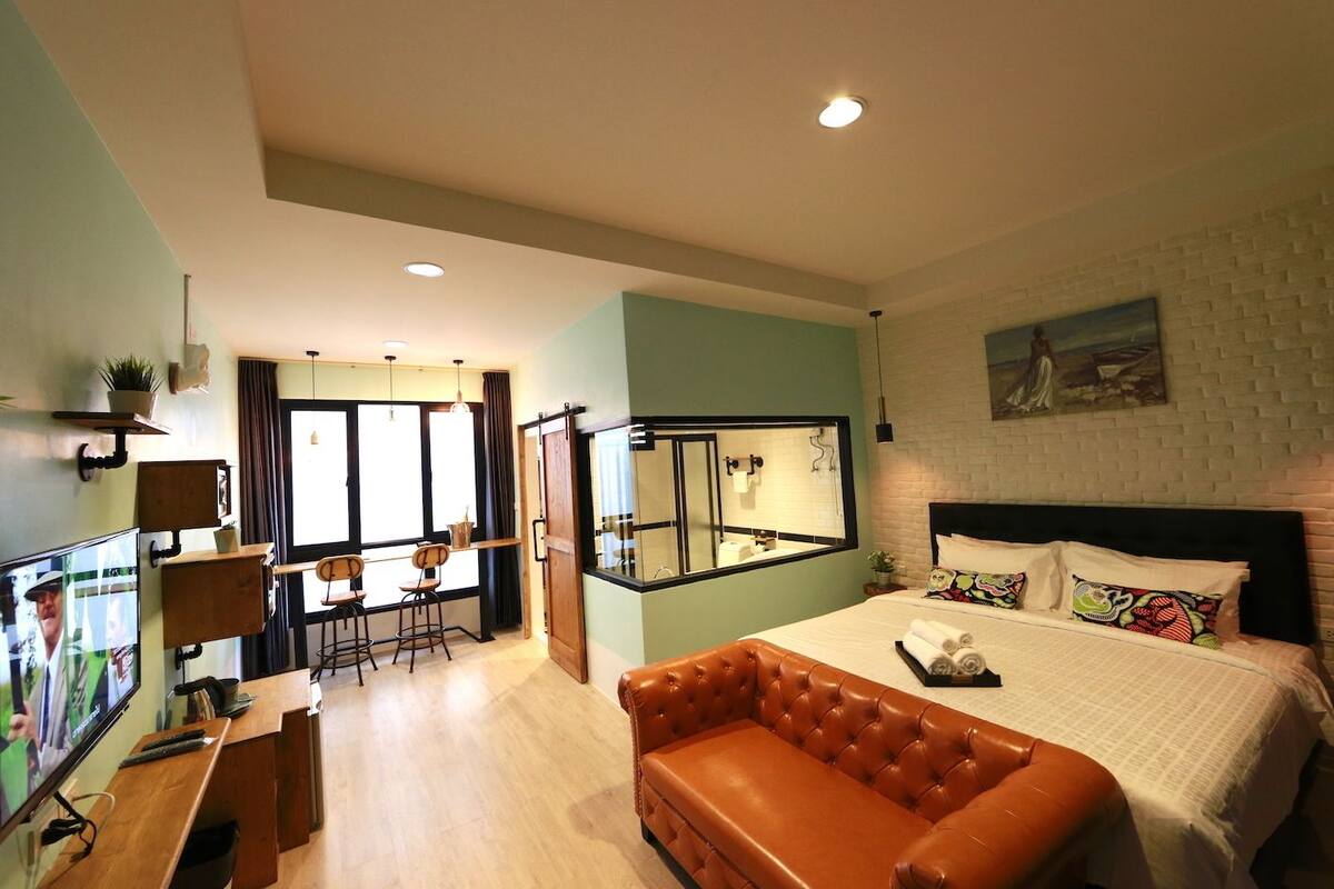 Une chambre d’hôtel moderne dotée d’un grand lit, d’un canapé en cuir, d’un coin repas avec une table et des chaises, et d’une grande fenêtre offrant une vue panoramique.