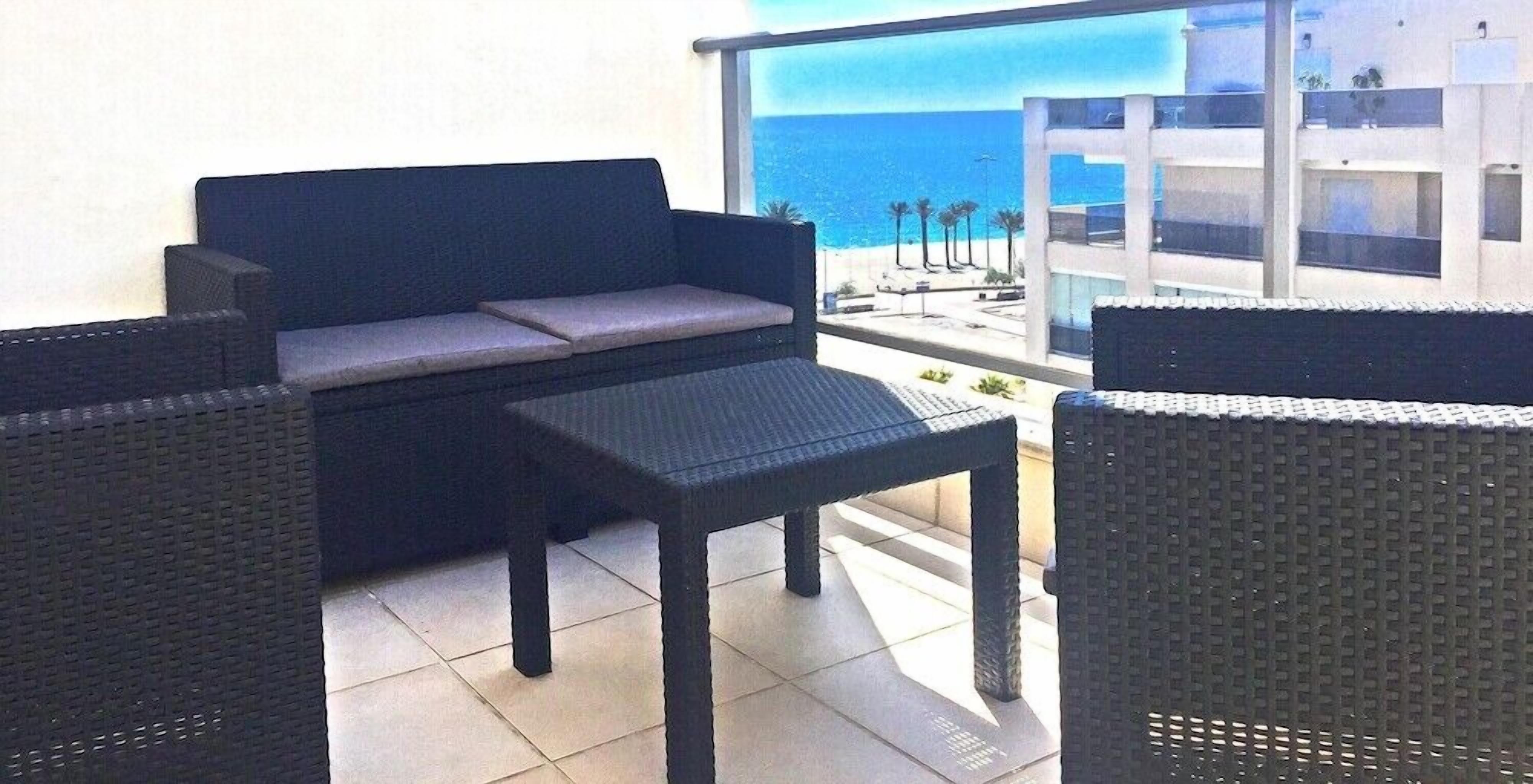 Ein Balkon mit einer dunkelgrauen Couch und zwei Korbstühlen, mit Blick auf einen Strand und ein modernes Gebäude.